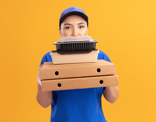 脸身穿蓝色制服 头戴鸭舌帽 拿着比萨饼盒和食品包的年轻送货员站在橙色的墙上 严肃地看着前方食物盒子女人