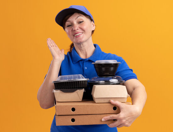 食物快乐的中年送货员 身穿蓝色制服 戴着帽子 手里拿着披萨盒和食品包 面带微笑地站在橙色的墙上 看着前面微笑女人拿着