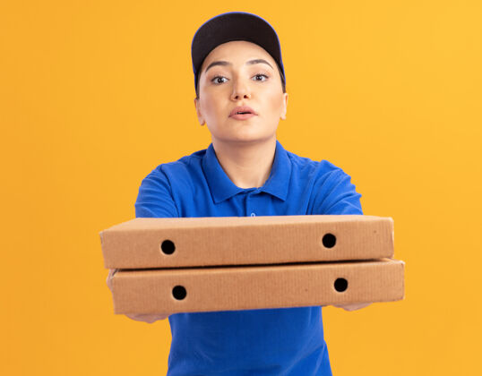 站着身着蓝色制服 戴着帽子 拿着披萨盒的年轻送货员站在橙色的墙上 自信地看着前方送货帽子披萨