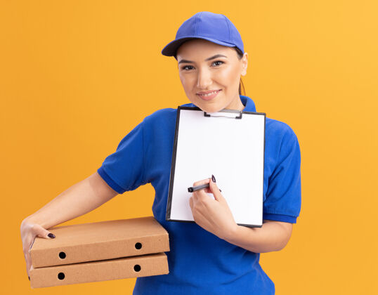 剪贴板身穿蓝色制服 头戴鸭舌帽的年轻女送货员拿着披萨盒和剪贴板 空白页上拿着铅笔要求签名 微笑着站在橙色的墙上看着前面铅笔签名帽子