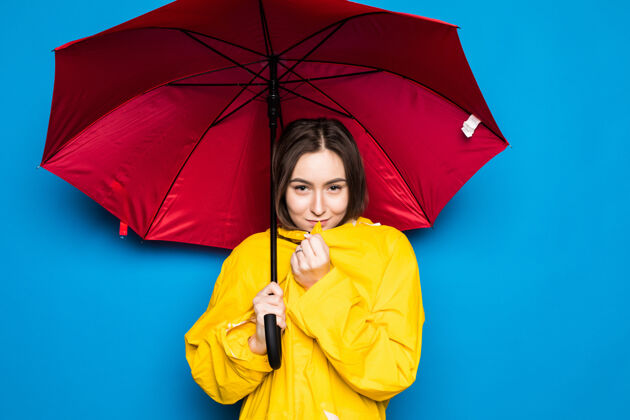 天气快乐的年轻女子拿着黄色雨衣和蓝色墙壁的雨伞下雨乐观脸