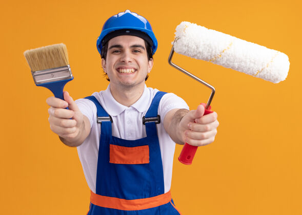 微笑快乐的年轻建筑工人身穿建筑制服 戴着安全帽 手里拿着油漆刷和压路机 站在橙色的墙上 面带微笑地看着前方年轻制服安全