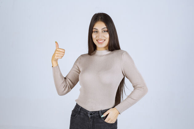 人类穿灰色毛衣的女孩在做竖起大拇指的招牌年轻姿势成功