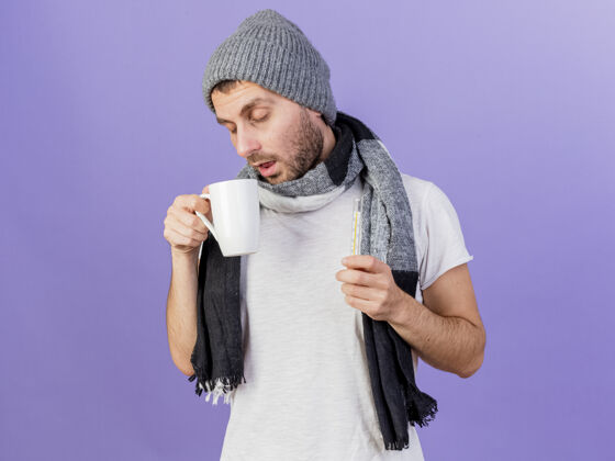 围巾闭着眼睛 戴着冬天的帽子 戴着围巾 手里拿着一杯茶和隔离在紫色背景上的温度计的年轻病人体温计闭着生病