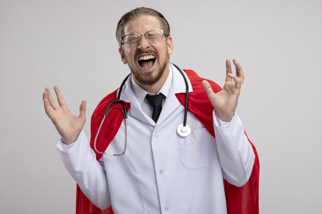 眼镜兴奋的年轻超级英雄 闭着眼睛 穿着带听诊器的医用长袍 戴着隔离在白色背景上的眼镜长袍年轻医疗