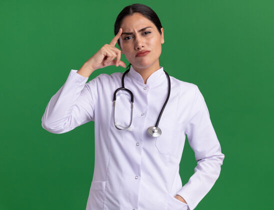 太阳穴年轻的女医生穿着医用外套 拿着听诊器 看起来很困惑 用食指指着她站在绿色墙壁上的太阳穴手指女人医生