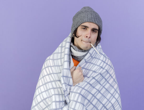 围巾虚弱的年轻病人戴着冬天的帽子 围巾裹着格子布 嘴里叼着温度计 隔离在紫色的背景上年轻人虚弱生病