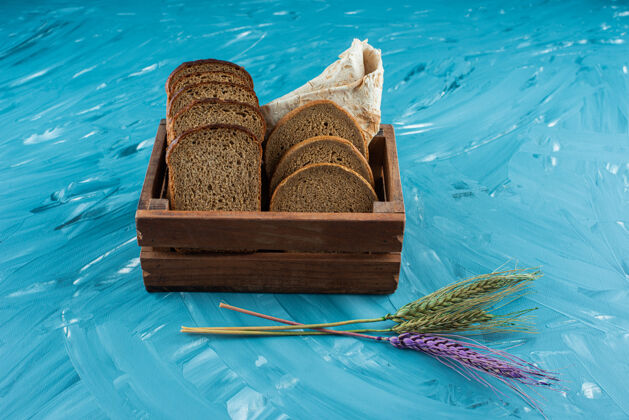 谷类食品一个木箱 里面装着蓝色背景的棕色新鲜面包片和小麦穗面包糕点盒子