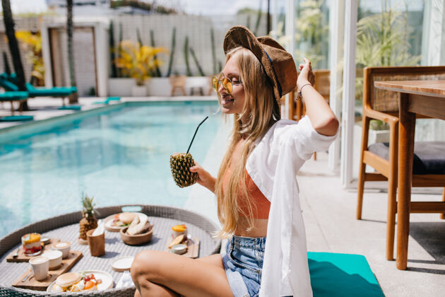 金发迷人的金发女人戴着黄色太阳镜 在游泳池边喝着可口的菠萝鸡尾酒梦幻般的女游客在度假胜地拍照时吃着水果微笑女性水