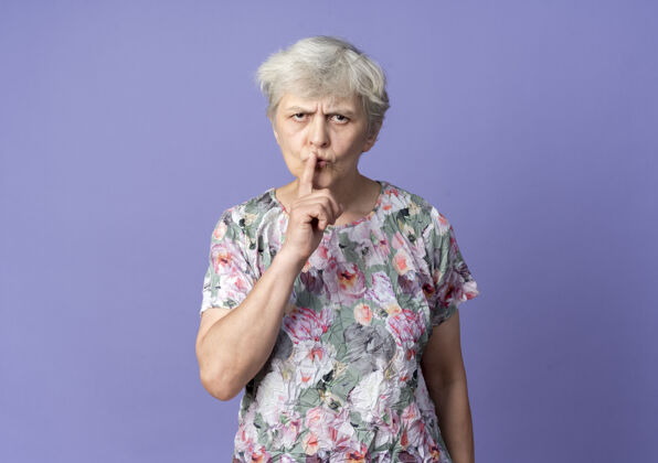 老人严肃的老妇人把手指放在嘴上做手势安静的标志孤立在紫色的墙上安静女人安静