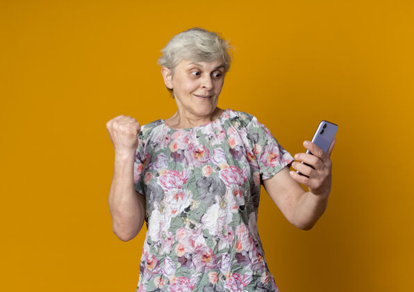 老人惊讶的老妇人举起拳头 拿着手机看着隔离在橙色墙上抱着举起拳头