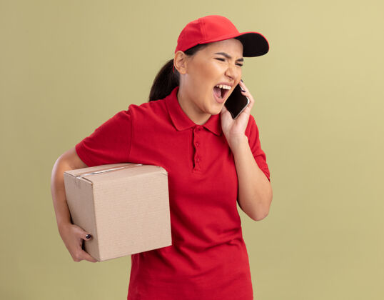 手机愤怒的年轻送货员身穿红色制服 戴着帽子 手里拿着纸板箱 站在绿色的墙上一边用手机说话一边大声喊叫纸板喊着站着