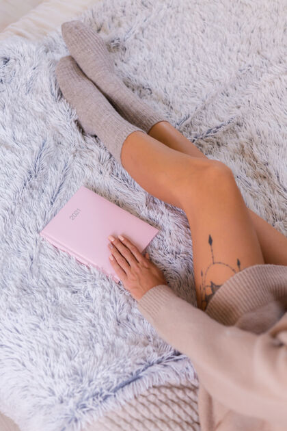 温暖穿着羊毛袜子和毛衣的女人 粉色笔记本上写着2021 臀部有大纹身女人坐在卧室的家里的床上计划者柔软坐着