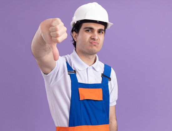 站起来年轻的建筑工人穿着建筑制服 戴着安全帽 不高兴地看着前面 竖起大拇指站在紫色的墙上年轻倒下建筑