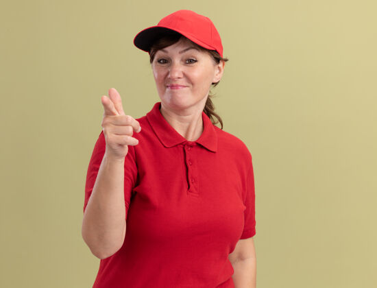 目录身穿红色制服 头戴鸭舌帽的中年女送货员自信地用食指指着前面的绿色墙壁制服信心交货