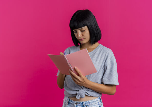 笔记本高兴的年轻黑发白种女人用钢笔写在笔记本上孤立的粉红色背景和复印空间年轻背景钢笔