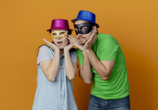 情侣年轻的夫妇惊讶地戴着粉色和蓝色的帽子戴上化装眼罩把手放在下巴上隔离在橙色的墙上化装舞会派对帽子