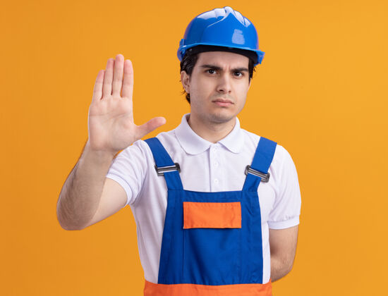 建筑年轻的建筑工人穿着建筑制服 戴着安全帽 严肃地看着前面 用手站在橘色的墙上做着停车的手势手手势停车