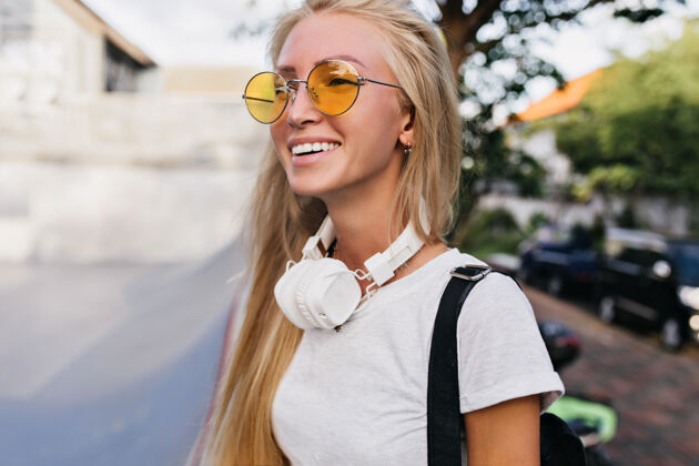 微笑脸上带着快乐表情的年轻女子戴着耳机在城市里走来走去戴着黄色太阳镜的金发美女在模糊的街道背景上笑着摆姿势金发人物城市