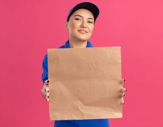 纸快乐的年轻送货员 身穿蓝色制服 戴着帽子 手里拿着纸包 站在粉红色的墙上 面带微笑地看着前面抱着包装年轻
