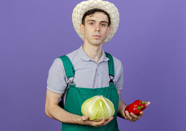花园自信的年轻男园丁戴着园艺帽 手里拿着卷心菜和辣椒空间园艺穿
