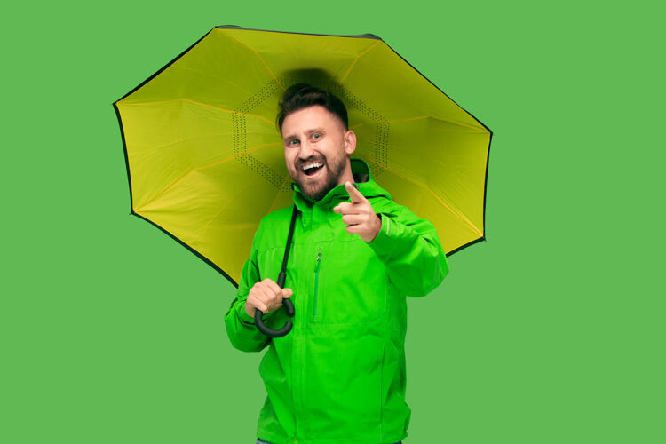 微笑帅气的胡须微笑的快乐的年轻人拿着雨伞 看着相机上的生动时尚的绿色工作室隔离秋天和寒冷的概念开始单独外套休闲