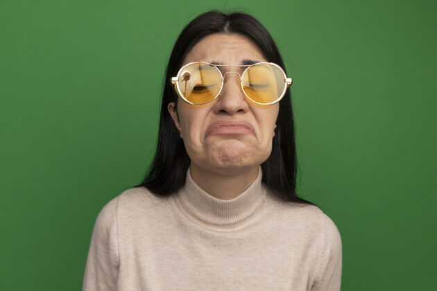 悲伤的一个戴着太阳眼镜的黑发白人女孩站在绿色的舞台上闭着眼睛封闭的成人的姿势的