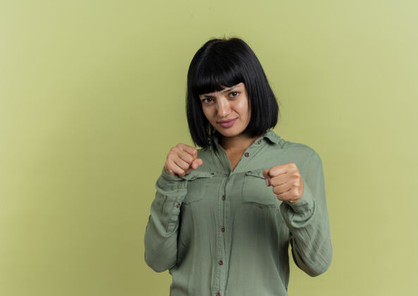 女人年轻的黑发白种女人保持拳头准备打孤立的橄榄绿背景与复制空间黑发拳打橄榄