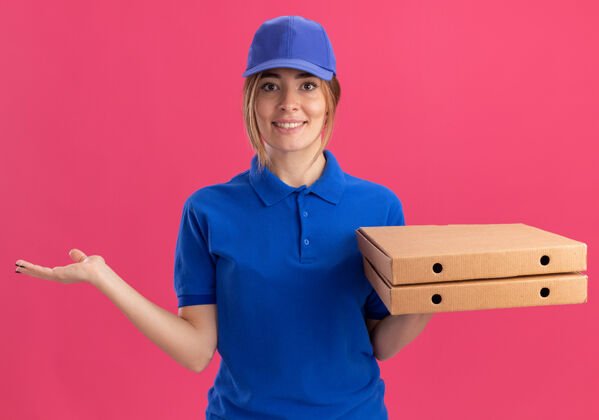 制服身着制服的年轻漂亮的送货女孩微笑着 手张开 手里拿着粉红色的比萨饼盒送货盒子披萨