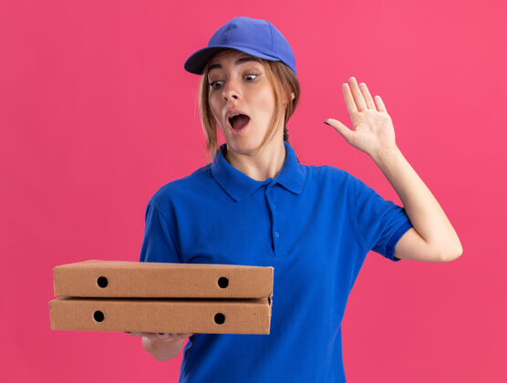 站穿着制服的年轻漂亮的送货女孩惊讶地举起手来 手里拿着粉红色的比萨饼盒姿势手制服