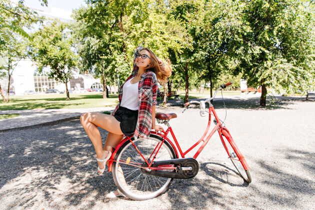 配件穿着格子衬衫的漂亮女孩在公园里享受夏天可爱的女模特坐在红色自行车上微笑的户外照片阳光漂亮驾驶