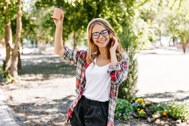 青少年一位戴着时髦眼镜的女士捧着一杯咖啡 笑着说在公园里散步的迷人的欧洲女孩的户外肖像自然卷发女人