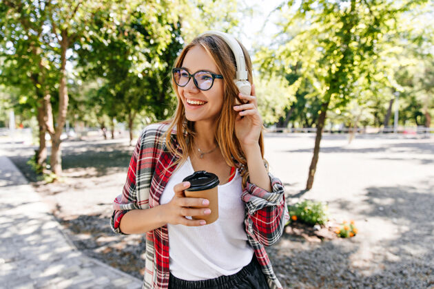 女人戴着眼镜戴着白色耳机摆姿势的金发女孩可爱的年轻女子端着茶站在大自然中的肖像女性微笑休息