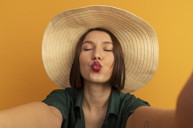 帽子戴着沙滩帽的漂亮白种女人假装亲吻相机在橙色上自拍自拍接吻沙滩