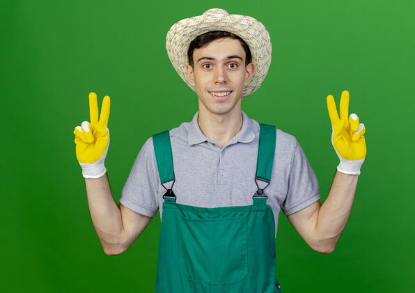 穿着微笑着的年轻男园丁戴着园艺帽子和手套 手势胜利手势 两只手在绿色背景上与复制空间隔离男性帽子园艺