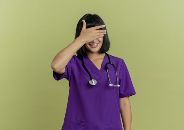 闭嘴微笑着的年轻黑发女医生穿着制服 手持听诊器闭上眼睛 手被隔离在橄榄绿背景上 留有复印空间医生听诊器女性