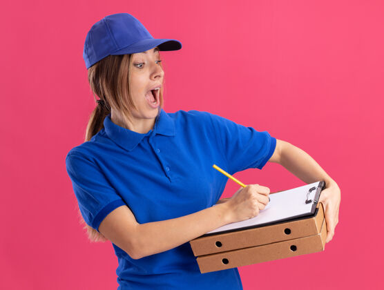 兴奋穿着制服的兴奋的年轻漂亮送货女孩拿着比萨饼盒 用粉红色的钢笔在剪贴板上写字盒子制服姿势
