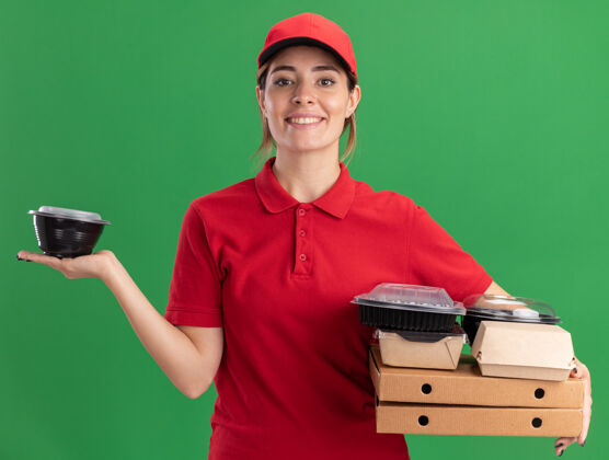 成人微笑着的年轻漂亮的送货女孩穿着制服拿着纸食品包装和比萨饼盒上的容器看着绿色的相机制服年轻人人
