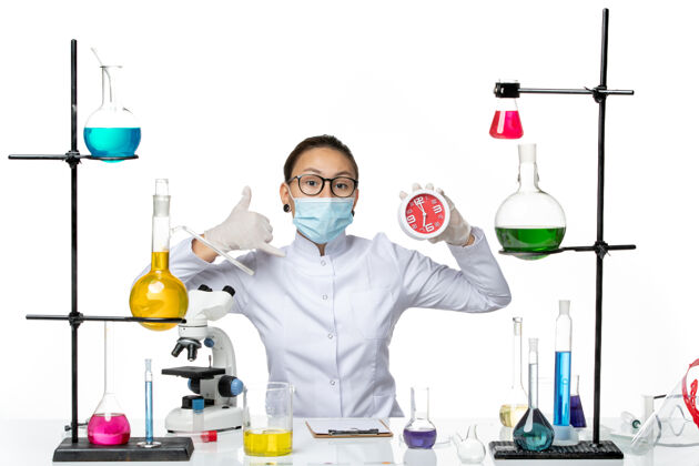 查看前视图穿着医疗服的女化学家 戴着面罩 在浅白色背景上拿着时钟病毒实验室化学covid-splash医生时钟医学