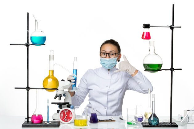 实验室前视图穿着医疗服的女化学家 戴着面罩 拿着浅白色背景上蓝色溶液的烧瓶 飞溅着病毒化学实验室的病毒医生面罩容器