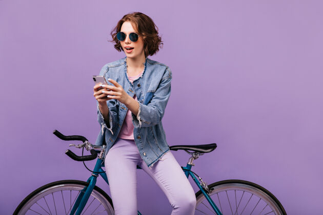 室内穿着休闲装的女人看着手机屏幕戴着墨镜的快乐白人女孩用智能手机和自行车乐趣寒冷快乐