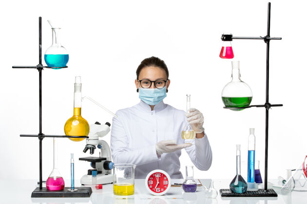 视图前视图穿着白色医疗服的女化学家 在桌子前面戴着面罩 白色背景上有溶液化学家实验室病毒冠状病毒飞溅前面桌子解决方案