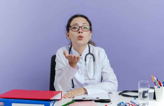 听诊器自信的年轻女医生穿着医用长袍 手持听诊器坐在办公桌旁 手持医疗工具 看着送出的口吻与世隔绝成人年轻紫色