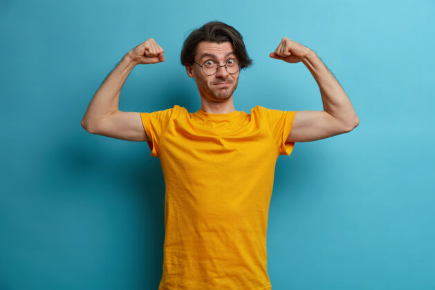 锻炼自信有力的男人举起双臂展示肌肉 展示定期锻炼的成果 穿着黄色t恤和眼镜 过着积极健康的生活方式 非常强壮保证二头肌休闲