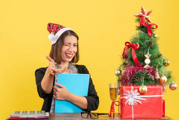 人风趣迷人的女士穿着西装 戴着圣诞老人的帽子和新年的装饰品 手里拿着文件 在黄色的办公室里与世隔绝圣诞老人圣诞老人成人