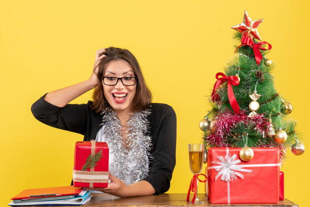 人一位情绪激动的商务女士穿着西装 戴着一副眼镜 展示着她的礼物 坐在一张桌子旁 桌子上放着一棵圣诞树眼镜封面女郎套装