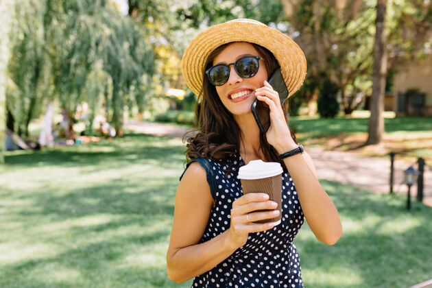休闲一个迷人的女人 戴着夏天的帽子 戴着黑色的太阳镜 穿着可爱的衣服 走在夏日公园里她一边喝着咖啡 一边讲着电话 情绪非常激动成人休闲阳光