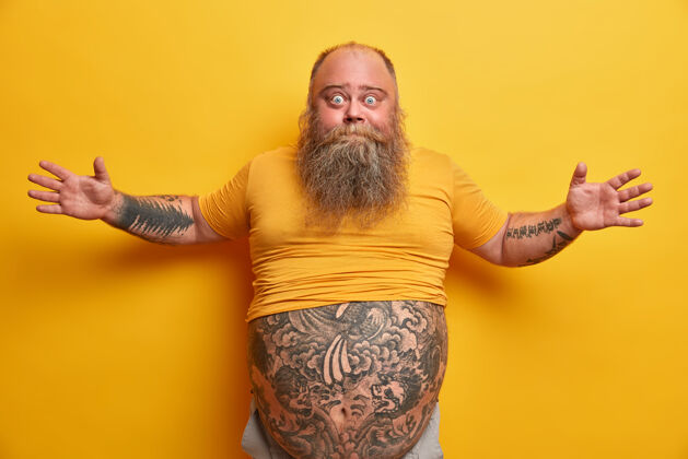 面部肥胖和不健康的生活方式的概念惊讶的眼男子展开双臂 讲述他看到的巨大的东西 手势积极 有纹身的身体和大肚子 隔离在黄色的墙壁上表情肥胖手掌