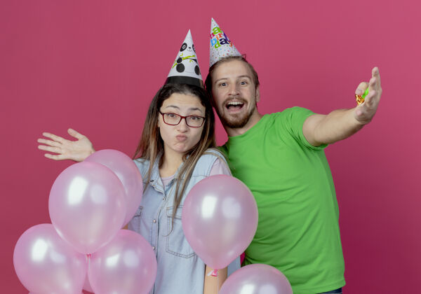 年轻快乐的年轻人戴着派对帽 惊讶的年轻女孩站在粉红色的墙上 氦气球被孤立气球粉色惊喜
