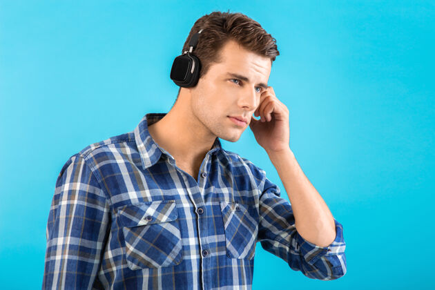 设备时尚帅气的年轻人 戴着无线耳机听音乐 玩得开心 现代风格 快乐的情绪 蓝色背景 穿着格子衬衫姿势脸时髦